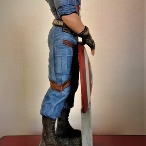 May include: A blue and red superhero figurine holding a red, white, and blue shield. The figurine is standing on a gray stone base.