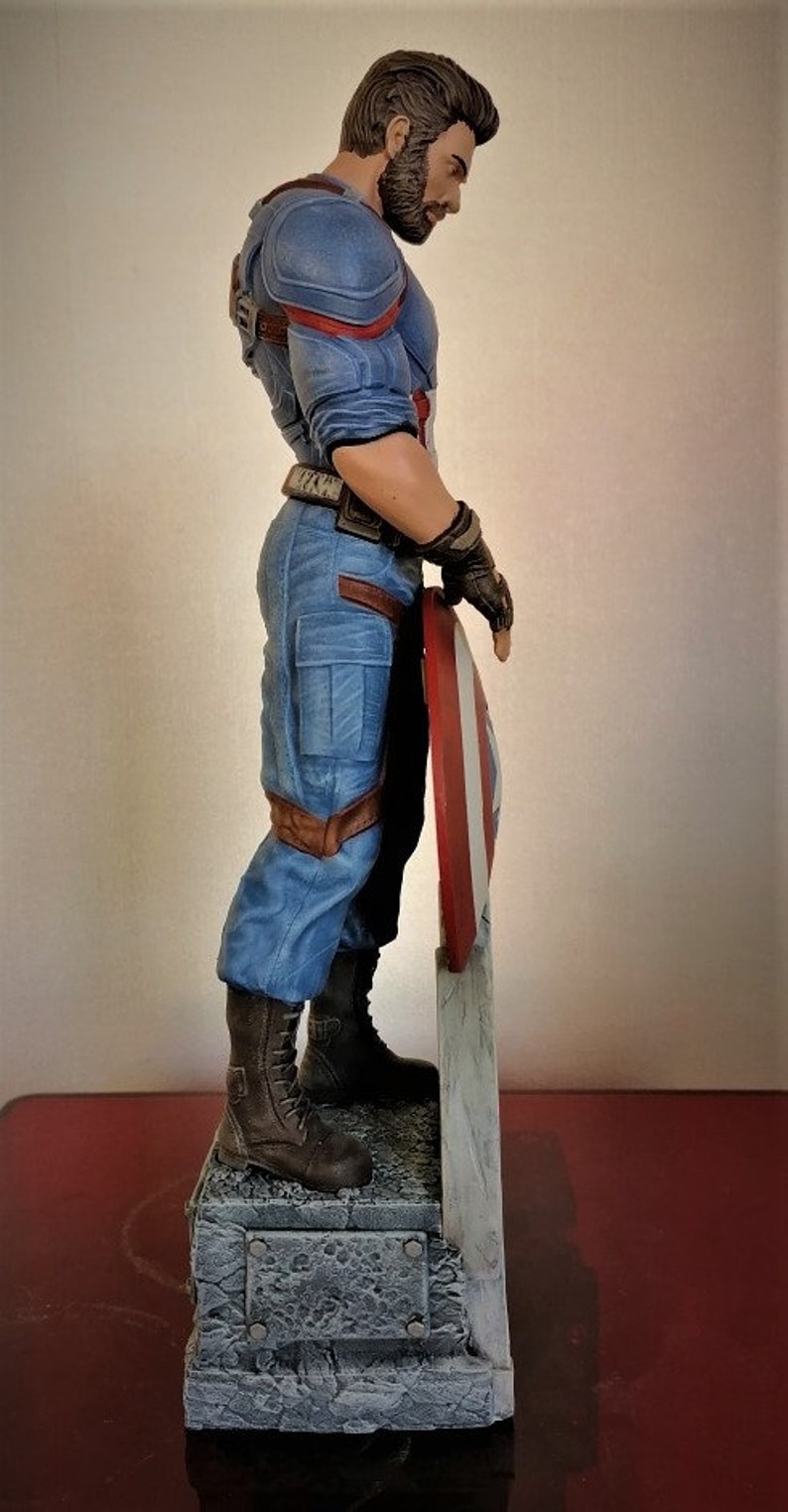 May include: A blue and red superhero figurine holding a red, white, and blue shield. The figurine is standing on a gray stone base.