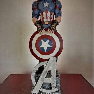 May include: A Captain America figurine holding his shield, standing on a base with the Avengers 'A' logo and the word 'LEGEND' inscribed on it.