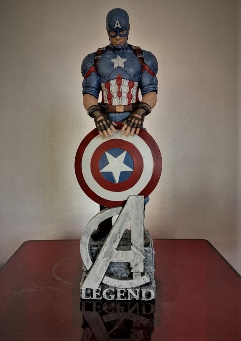 May include: A Captain America figurine holding his shield, standing on a base with the Avengers 'A' logo and the word 'LEGEND' inscribed on it.