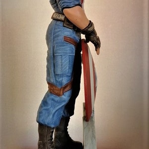 May include: A blue and red action figure of Captain America standing on a gray stone base. He is holding his shield in his right hand and is wearing a blue and red costume with a red, white, and blue helmet.
