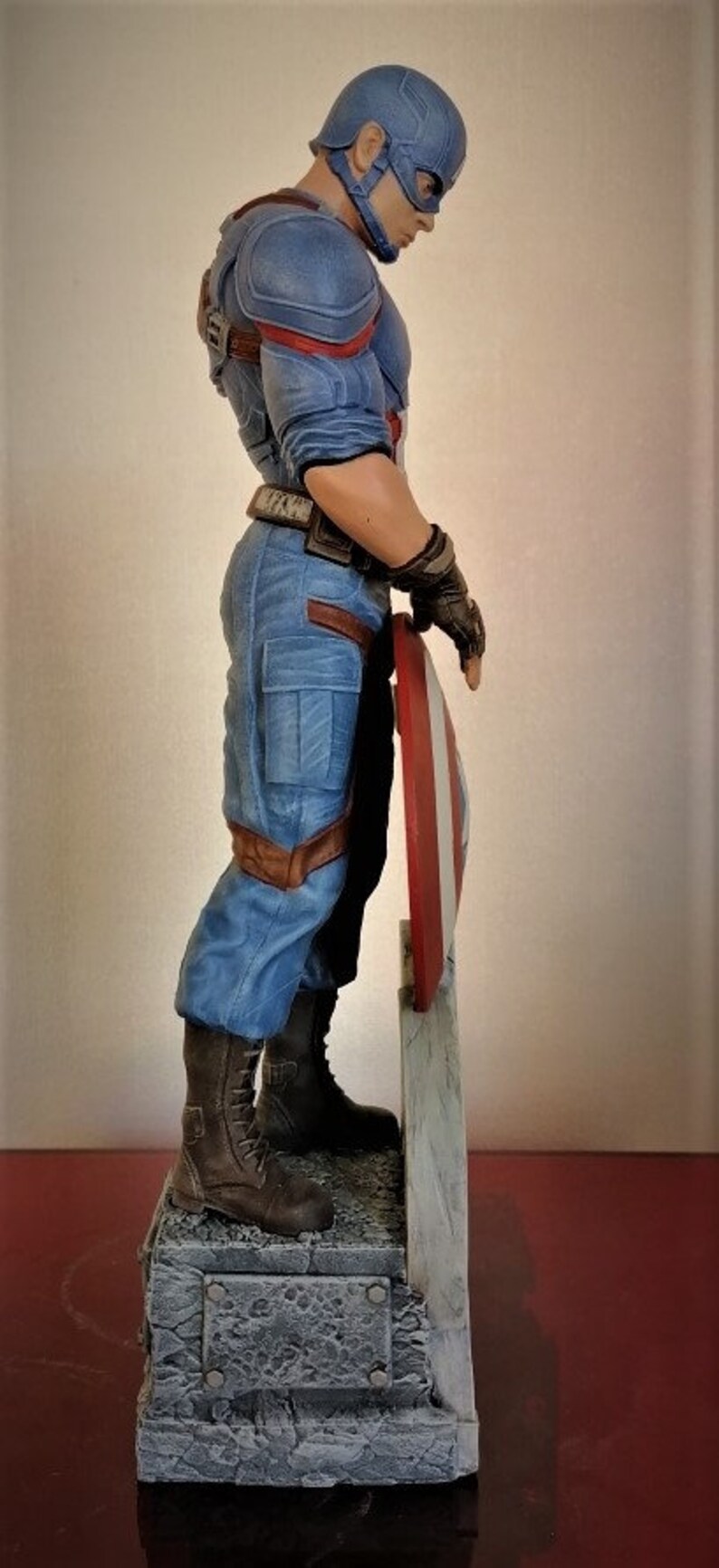 May include: A blue and red action figure of Captain America standing on a gray stone base. He is holding his shield in his right hand and is wearing a blue and red costume with a red, white, and blue helmet.
