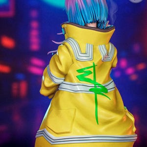 May include: A figurine of a character with blue and pink hair, wearing a yellow jacket with a green design on the back. The jacket has white and silver accents. The figure is set against a blurred, dark background with neon lights. The text "Peach Figure" is visible.