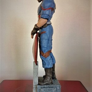 May include: A detailed figurine of Captain America in his blue and red costume, holding his shield. The figurine is standing on a stone base.