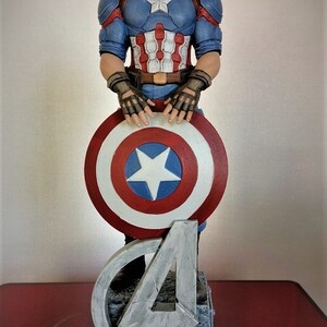 May include: A Captain America action figure with a red, white, and blue costume, holding his shield. The figure is standing on a base with the Avengers logo and the word "LEGEND" printed on it.