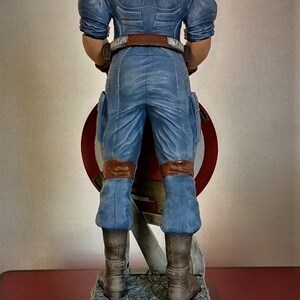 May include: A blue and red Captain America action figure stands on a gray stone base. The figure is holding a red, white, and blue shield in front of it. The figure is wearing a blue and red costume with a brown belt and brown boots.