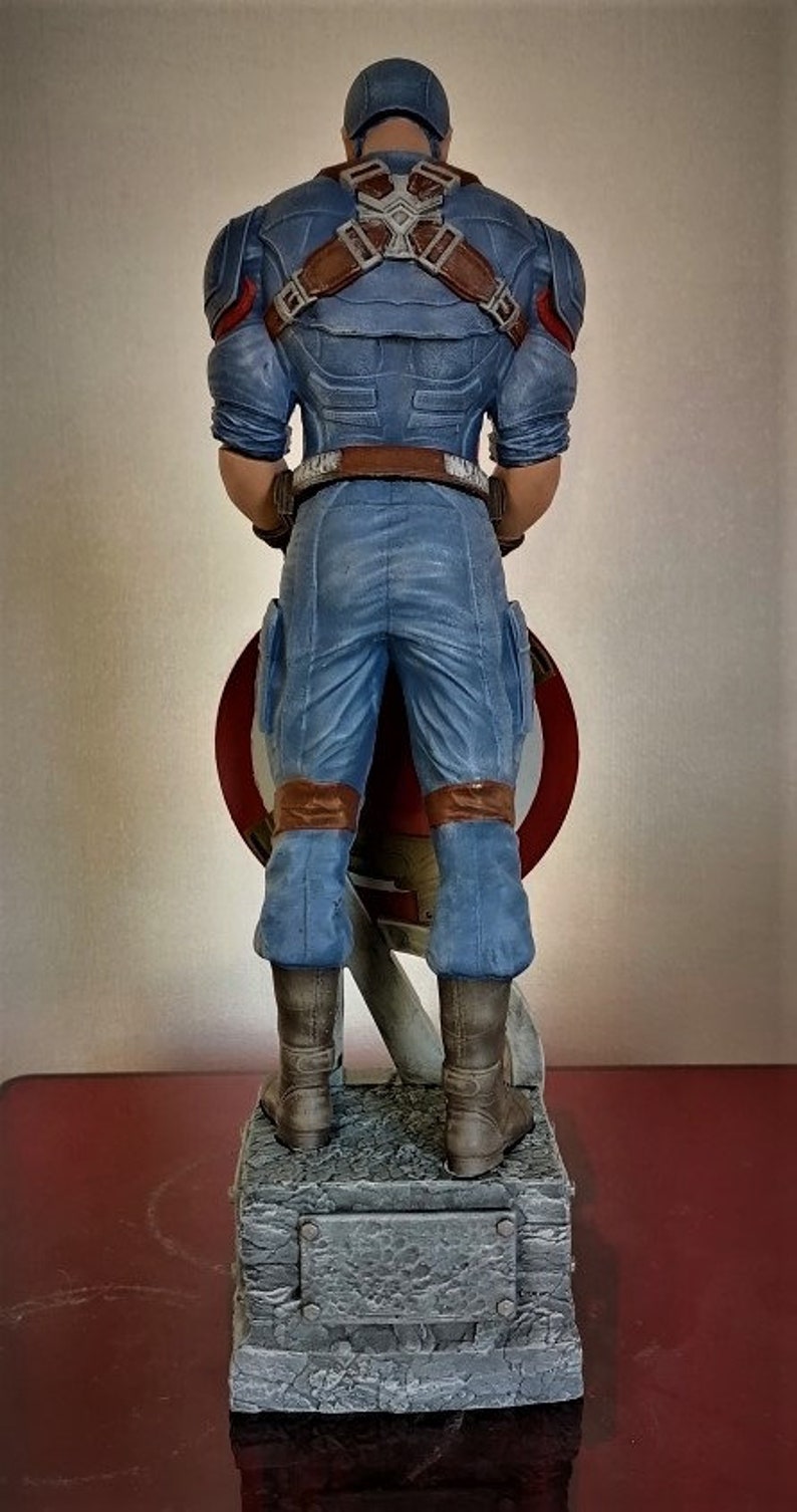 May include: A blue and red Captain America action figure stands on a gray stone base. The figure is holding a red, white, and blue shield in front of it. The figure is wearing a blue and red costume with a brown belt and brown boots.