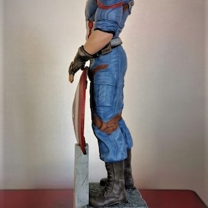 May include: A blue and red Captain America action figure stands on a gray stone base, holding his shield in his left hand. The figure is wearing a blue and red costume with a red star on his chest. The figure is posed in a heroic stance, with his right leg forward and his left leg back.