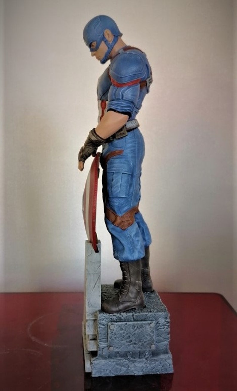 May include: A blue and red Captain America action figure stands on a gray stone base, holding his shield in his left hand. The figure is wearing a blue and red costume with a red star on his chest. The figure is posed in a heroic stance, with his right leg forward and his left leg back.