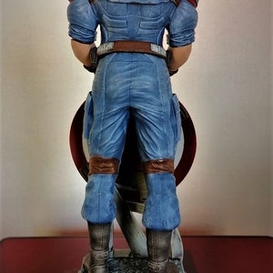 May include: A blue and brown action figure of Captain America standing on a gray base. The figure is holding a red, white, and blue shield.