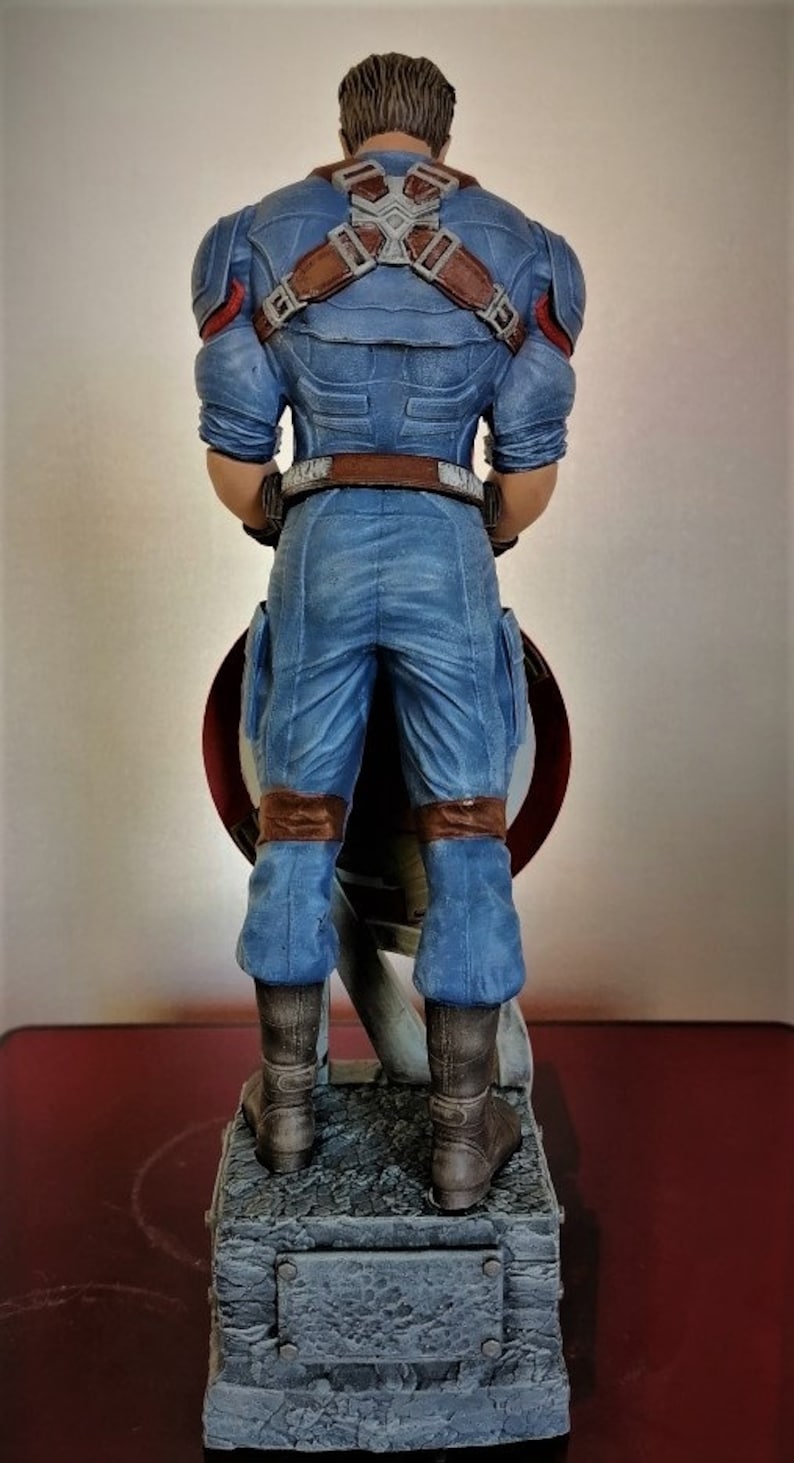 May include: A blue and brown action figure of Captain America standing on a gray base. The figure is holding a red, white, and blue shield.