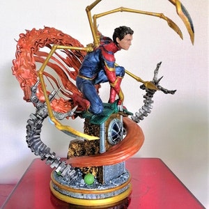 May include: A detailed action figure of Spider-Man in his red and blue suit with gold accents, standing on a base with a web-like design. The figure has a dynamic pose with one arm outstretched and the other holding a web shooter. The figure is surrounded by a fiery orange web.