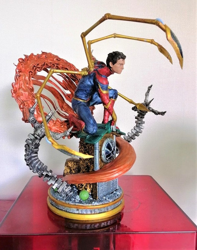 May include: A detailed action figure of Spider-Man in his red and blue suit with gold accents, standing on a base with a web-like design. The figure has a dynamic pose with one arm outstretched and the other holding a web shooter. The figure is surrounded by a fiery orange web.