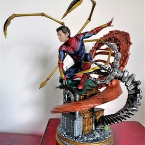 May include: A red and blue action figure of Spider-Man with gold accents, standing on a detailed base with a mechanical arm and a red and gold web. The figure is posed in a dynamic action pose.
