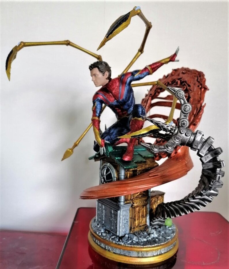 May include: A red and blue action figure of Spider-Man with gold accents, standing on a detailed base with a mechanical arm and a red and gold web. The figure is posed in a dynamic action pose.