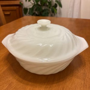 May include: A white, lidded casserole dish with a spiral design. The dish has two handles and a small knob on the lid. The dish is made of a glossy, opaque material.