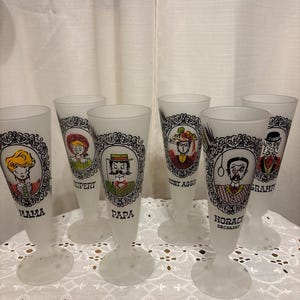 May include: Set of six frosted glass champagne flutes. Each glass features a different cartoon character portrait in a black and white ornate frame. The names "Mama", "Rupert", "Papa", "Aunt Aggie", "Horace Deceased", and "Grampa" are printed on the glasses.