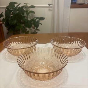May include: Three vintage, light amber-colored glass bowls. Each bowl has a ribbed exterior and a detailed, patterned interior. The bowls are arranged on a white cloth, with a wooden table and a green plant in the background.