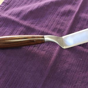 May include: A stainless steel spatula with a rectangular blade and a dark wood handle. The spatula is lying on a purple cloth.