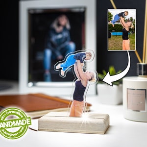 Könnte beinhalten: Ein transparenter Acryl-Bilderrahmen mit einer Holzbasis. Der Rahmen zeigt ein Foto einer Frau, die ein Kind in die Luft hält. Das Foto ist auf dem Acryl gedruckt und von beiden Seiten sichtbar.