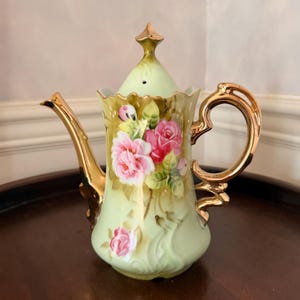 May include: A vintage porcelain teapot with a light green base and gold accents. The teapot features a floral design with pink roses and green leaves. The handle and spout are gold-colored, and the lid is topped with a gold finial.