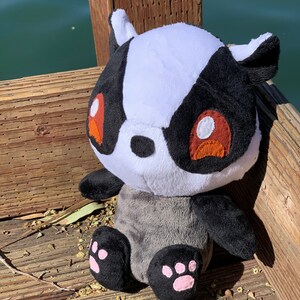 May include: A plush toy badger with black and white fur, red eyes, and pink paw pads. The badger is sitting on a wooden surface.