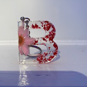 May include: A clear resin keychain shaped like the letter B with a pink flower pressed inside. The resin is filled with red glitter.