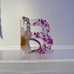 May include: A clear resin keychain shaped like the letter "B" with a pressed flower inside. The resin is filled with purple glitter.