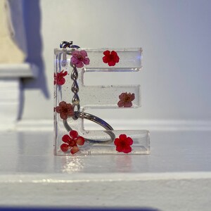 May include: Clear resin letter "E" keychain with embedded red and pink dried flowers. The keychain has a silver-colored chain and keyring. The letter is approximately 2 inches tall and sits on a white surface.