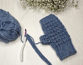 May include: A blue crocheted fingerless glove with a matching ball of yarn and a crochet hook. The glove is partially completed, showing the textured stitch pattern. The yarn is a soft, muted blue color. The hook has a white handle.