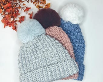 May include: Four knitted winter hats in shades of gray, pink, and blue, each topped with a fluffy pom-pom. The hats are arranged in a diagonal stack, showcasing their textured knit design. A sprig of orange leaves adds a seasonal touch.