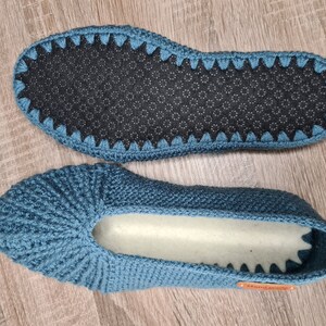 May include: Two blue crochet slippers. The soles are dark gray with a non-slip pattern. The slippers are handmade, as indicated by a small orange tag. The top of the slippers are a solid blue color with a ribbed design.