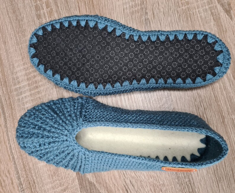 May include: Two blue crochet slippers. The soles are dark gray with a non-slip pattern. The slippers are handmade, as indicated by a small orange tag. The top of the slippers are a solid blue color with a ribbed design.