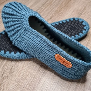 May include: A pair of teal crochet slippers with a textured design. The slippers have a non-slip sole and a small leather tag that reads "Hand made with Love". The slippers are designed for indoor use.