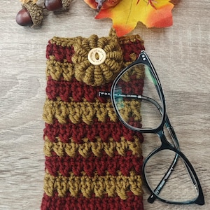 May include: A hand-crocheted glasses case in burgundy and olive green, with a textured stitch design. The case has a matching crochet flower and a wooden button fastening. A pair of black-framed glasses is placed next to the case.