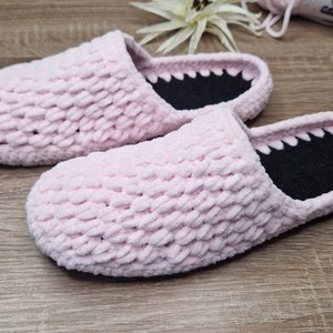 Op de afbeelding: Een paar lichtroze, zachte pantoffels. De pantoffels hebben een getextureerde, gebreide uitstraling en een zwart interieur. De zolen zijn donker en de pantoffels liggen op een houten oppervlak. De pantoffels zijn ontworpen voor gebruik binnenshuis.
