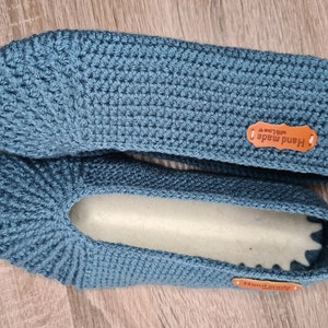May include: A pair of handmade, teal blue crochet slippers. The slippers have a textured stitch pattern and a soft, light-colored interior. Each slipper has a small, brown leather tag that reads "Hand made with love".