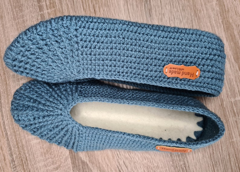 May include: A pair of handmade, teal blue crochet slippers. The slippers have a textured stitch pattern and a soft, light-colored interior. Each slipper has a small, brown leather tag that reads "Hand made with love".