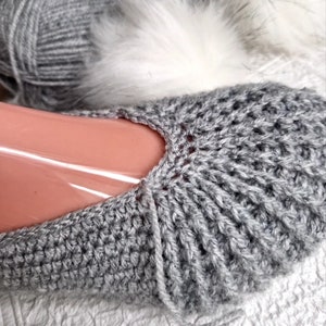 May include: Close-up of a gray crocheted slipper. The slipper is made of a ribbed knit pattern and is partially covering a foot form. A ball of gray yarn and a white fluffy material are in the background.