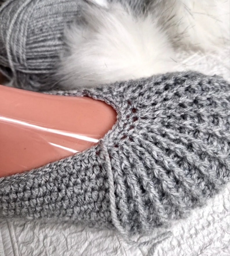 May include: Close-up of a gray crocheted slipper. The slipper is made of a ribbed knit pattern and is partially covering a foot form. A ball of gray yarn and a white fluffy material are in the background.