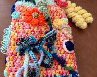 May include: A rectangular crochet cover featuring a vibrant mix of pink, orange, yellow, and blue yarn. The surface is decorated with three-dimensional crocheted elements including a bright orange flower, a small ice cream cone, and yellow spirals.