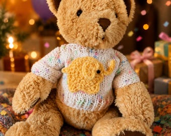 May include: A plush tan teddy bear with dark brown eyes and nose, wearing a white knitted jumper featuring a textured yellow elephant appliqué on the chest. The bear has soft, fuzzy fur and dark brown fabric patches on the soles of its feet.