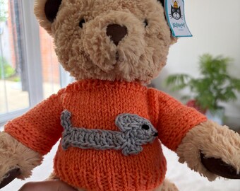 May include: A plush teddy bear with light brown fur wearing an orange knitted jumper. The jumper features a grey crocheted dachshund dog appliqué on the front. A blue Jellycat brand tag is attached to the bear's ear, displaying a cat logo.