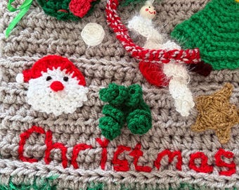 May include: A beige crochet wall hanging featuring the red stitched text Christmas. Embellishments include a green wreath with a red bow, a Santa Claus face, a gold star, a green tree, and a small white figure with a red and white scarf.