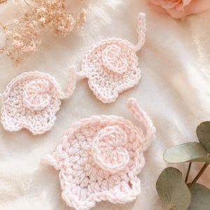 May include: Three light pink crochet elephant appliqués, each featuring a textured body, a small curled trunk, and a rounded ear. These handmade yarn pieces are crafted with a consistent stitch pattern and are arranged in a group.