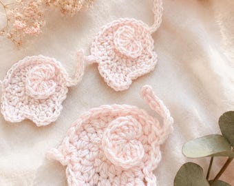 May include: Three light pink crochet elephant appliqués, each featuring a textured body, a small curled trunk, and a rounded ear. These handmade yarn pieces are crafted with a consistent stitch pattern and are arranged in a group.