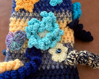 May include: A rectangular crochet pouch featuring horizontal stripes in navy blue, light blue, and yellow. The surface is embellished with various three-dimensional crochet appliqués, including a yellow star, light blue flowers, a small evil eye bead, and dark blue curly tendrils.