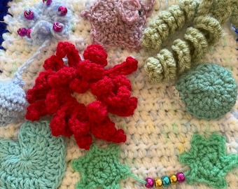 May include: A square crochet sensory blanket featuring a white base with multicoloured flecks and a dark blue border. It is decorated with various crocheted shapes including a pink elephant, a red flower, green spirals, a light blue heart, and beads.