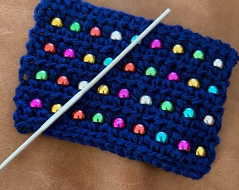 May include: A rectangular piece of dark blue crocheted fabric featuring rows of small, shiny, multi-coloured metallic beads in shades of red, green, yellow, pink, and silver. A long, thin, light-coloured crochet hook is placed diagonally across the fabric.