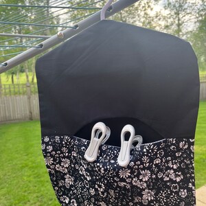 Puede incluir: Una bolsa de lavander&iacute;a de tela negra con estampado floral y un gancho blanco. La bolsa tiene un bolsillo en la parte delantera para guardar pinzas para la ropa. Hay tres pinzas para la ropa blancas en el bolsillo.